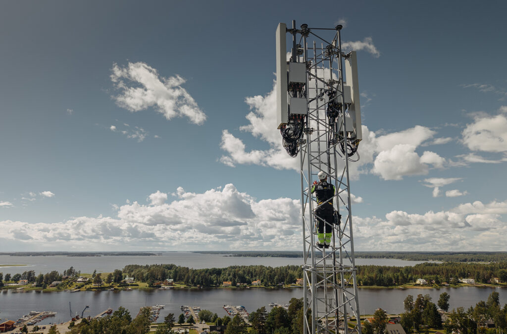Flygfoto av en tekniker som klättrar högt upp i en fackverksmast med antennpaneler, med en sjö och skog synlig i bakgrunden.