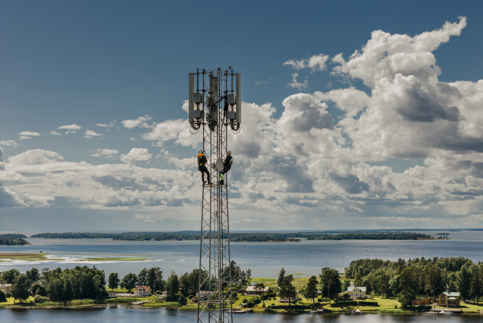 Flygfoto på avstånd av en hög mast med två tekniker som klättrar nära toppen, omgiven av en skärgård med öar och vatten.