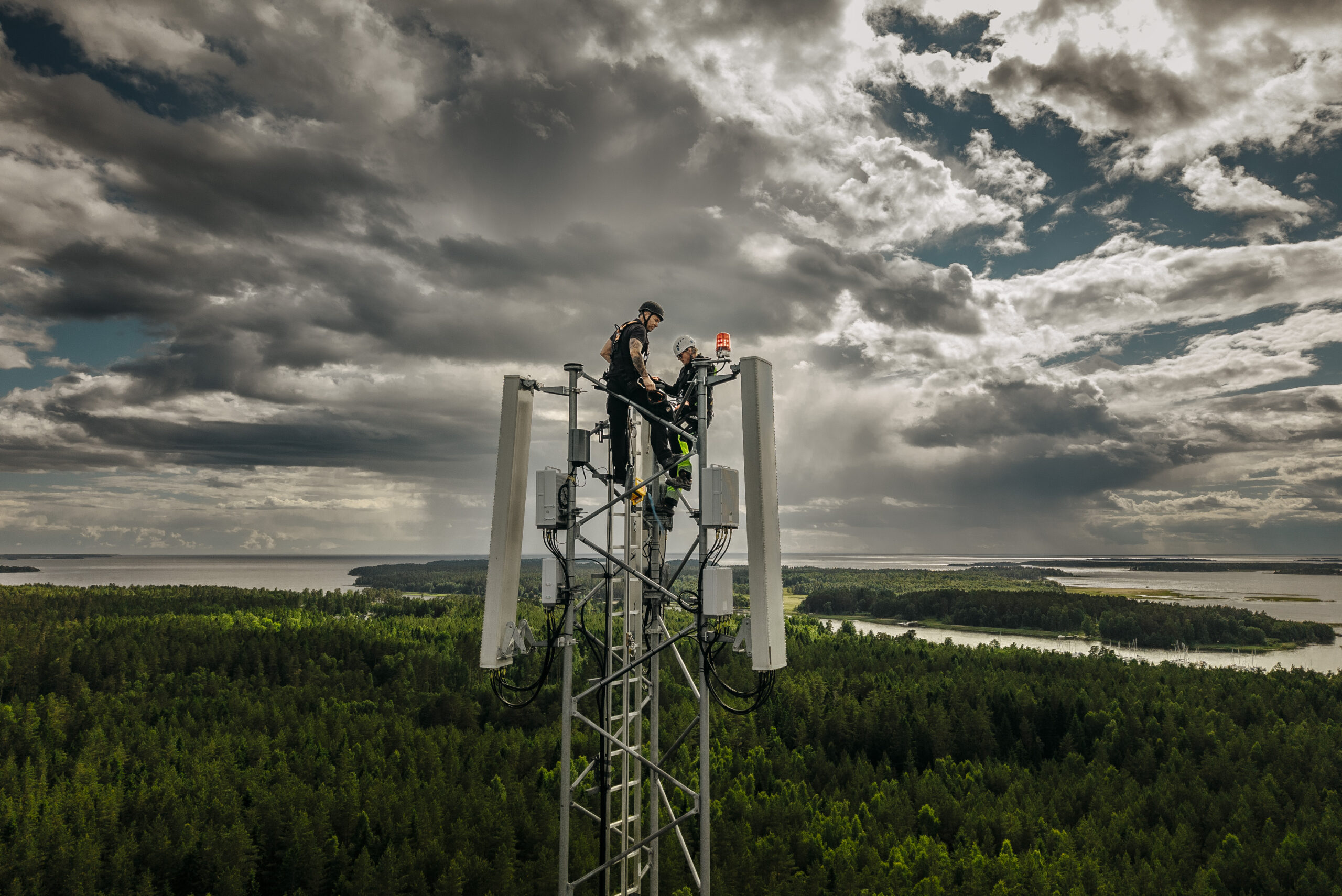 Flygfoto av två tekniker som arbetar högst upp på en mast ovanför ett vidsträckt skogs- och sjölandskap under en dramatisk molnig himmel.