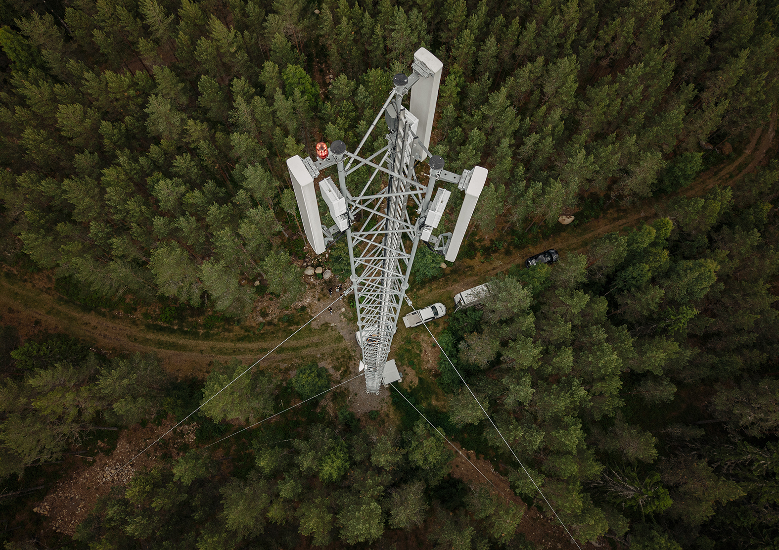 Flygfoto rakt uppifrån av en mast med antennpaneler omgiven av tät skog, med en skogsväg och parkerade servicebilar nedanför.