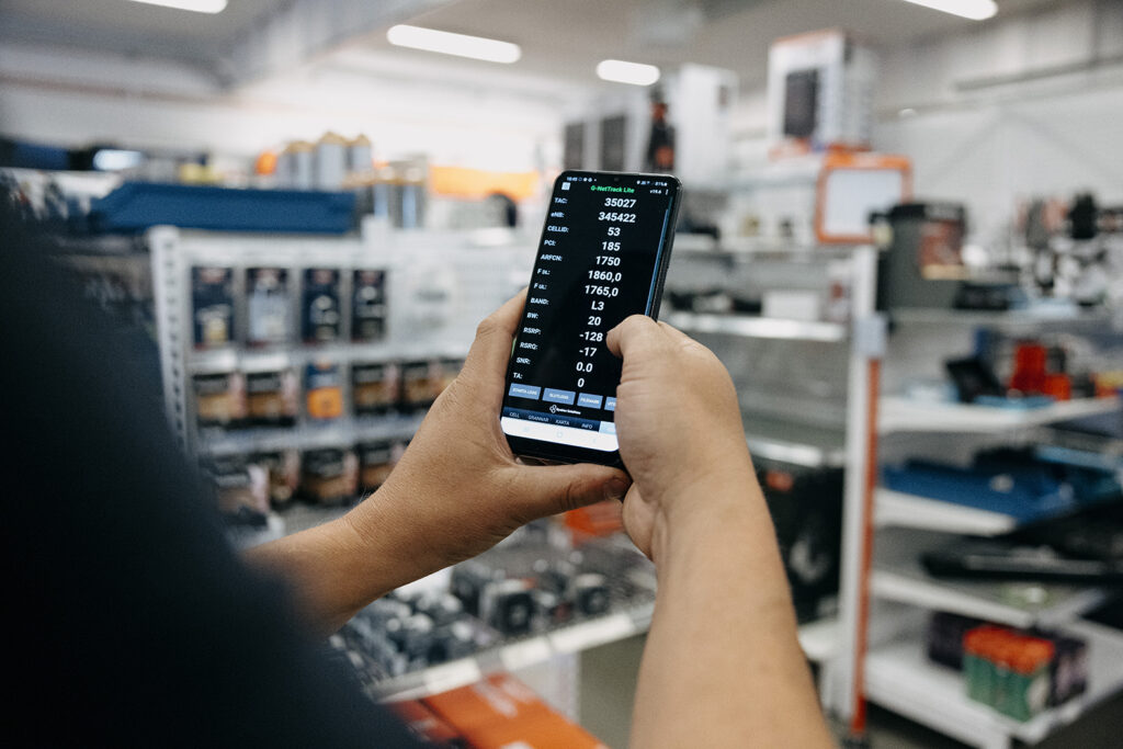 En person håller en mobiltelefon med en mätapp som visar nätverksdata i ett lager med hyllor av utrustning.