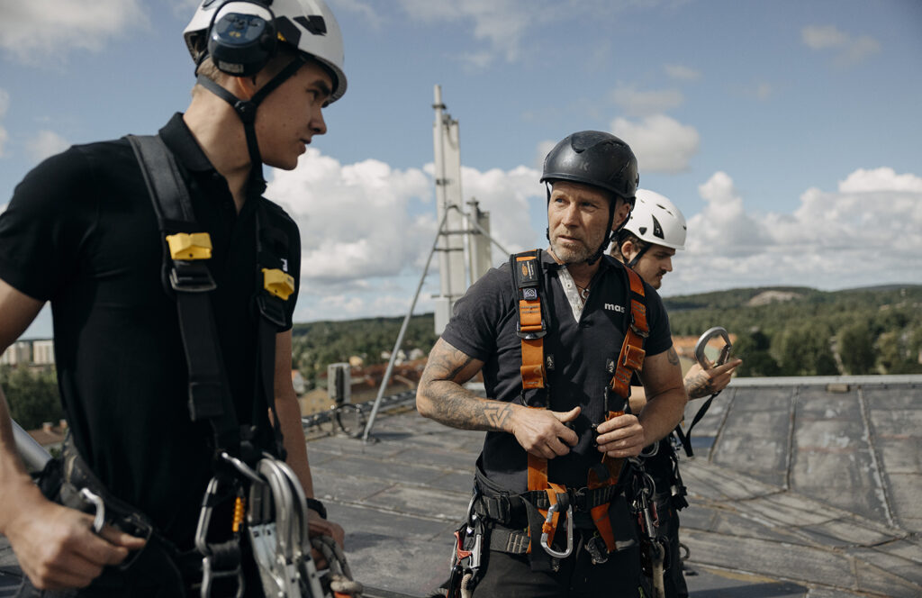 Tre tekniker i klätterutrustning och hjälmar samtalar på ett tak med en mast och en stad synlig i bakgrunden.
