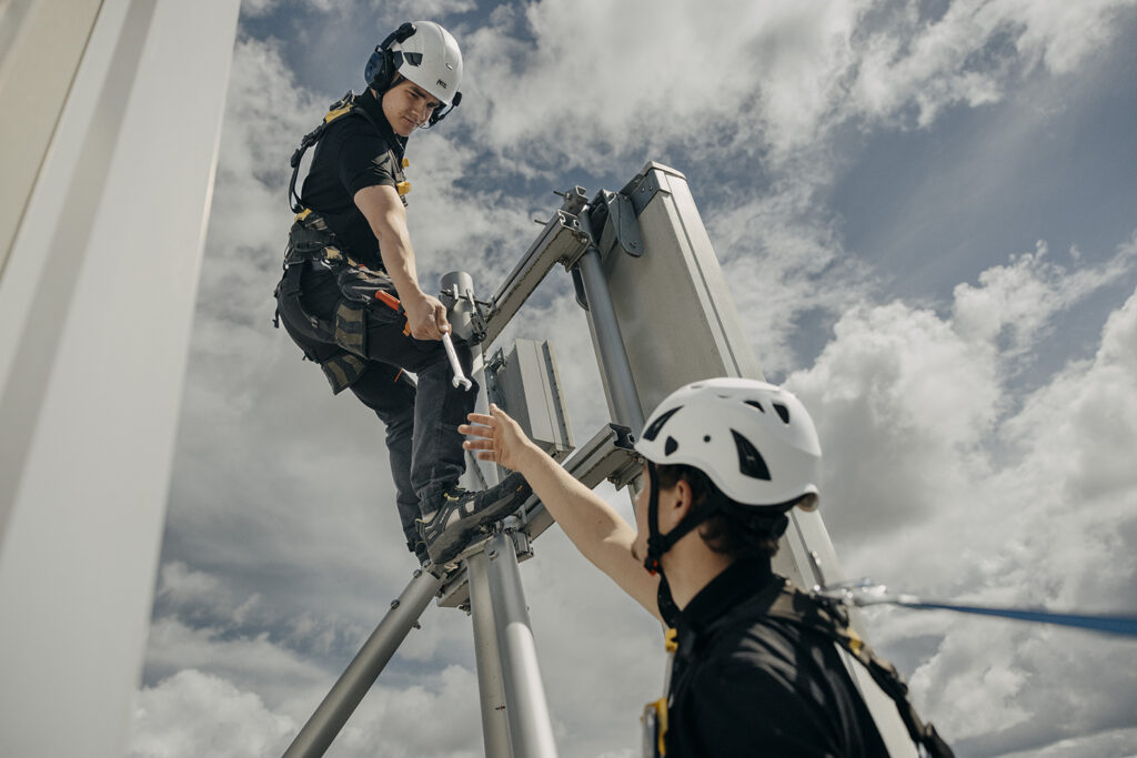 En tekniker med hjälm sträcker sig fram med en skiftnyckel mot en antennpanel medan en kollega längre ner räcker upp handen, fotograferat underifrån mot en molnig himmel.