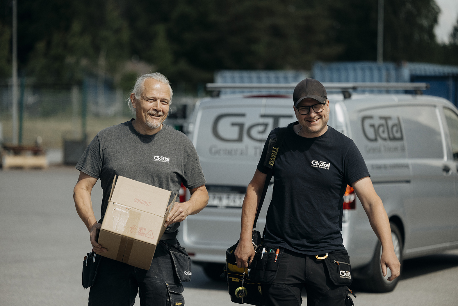 Två leende män i GeTel-tröjor bär en kartong och en verktygsväska framför en skåpbil märkt GeTel.