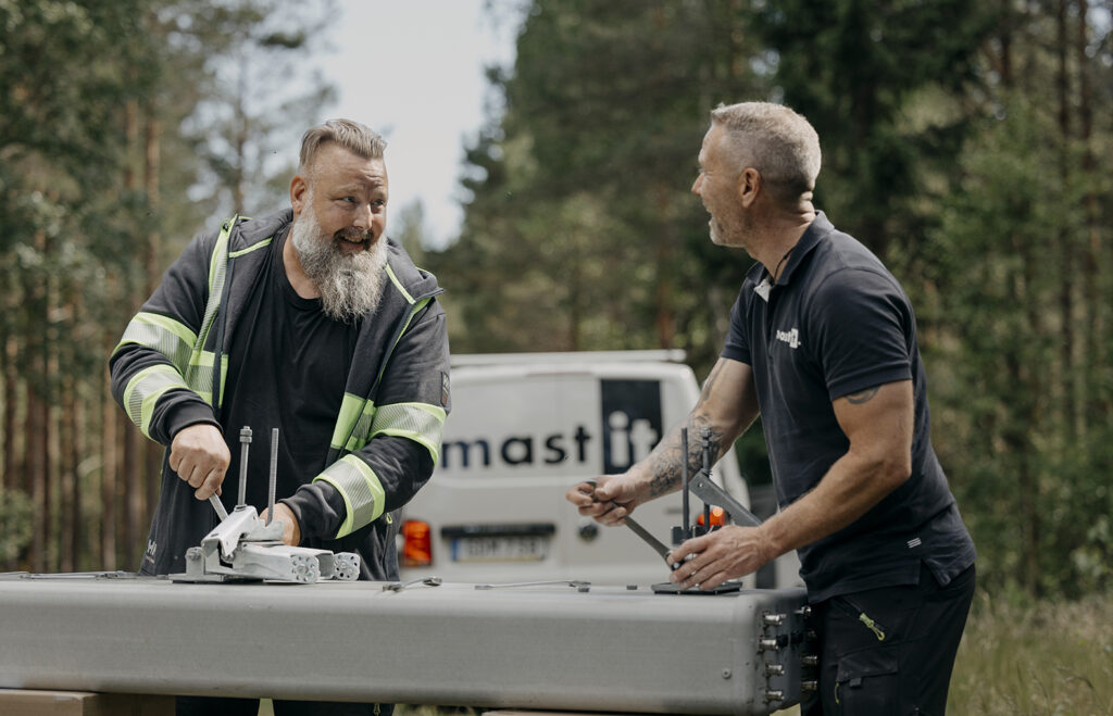 Två tekniker samtalar och arbetar med metallkomponenter på flaket av en skåpbil märkt mastIT i skogen.