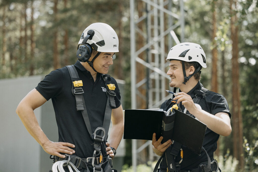 Två leende tekniker med hjälmar samtalar utomhus, en av dem skriver på en surfplatta i ett skyddsfodral med en mast synlig i bakgrunden.