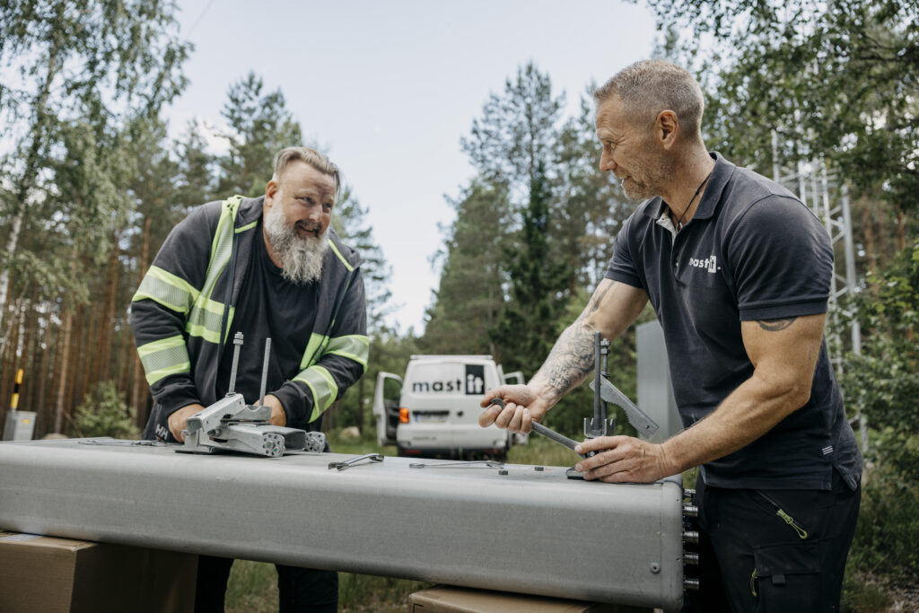 Två tekniker utomhus i skogen böjer sig över en lång grå utrustningslåda på ett bord och skruvar fast komponenter, med en skåpbil märkt mastIT synlig i bakgrunden.