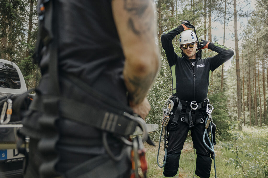 En leende tekniker med hjälm och solglasögon justerar sin klättersele i en skog, fotograferad förbi en kollegas tatuerade arm i förgrunden.