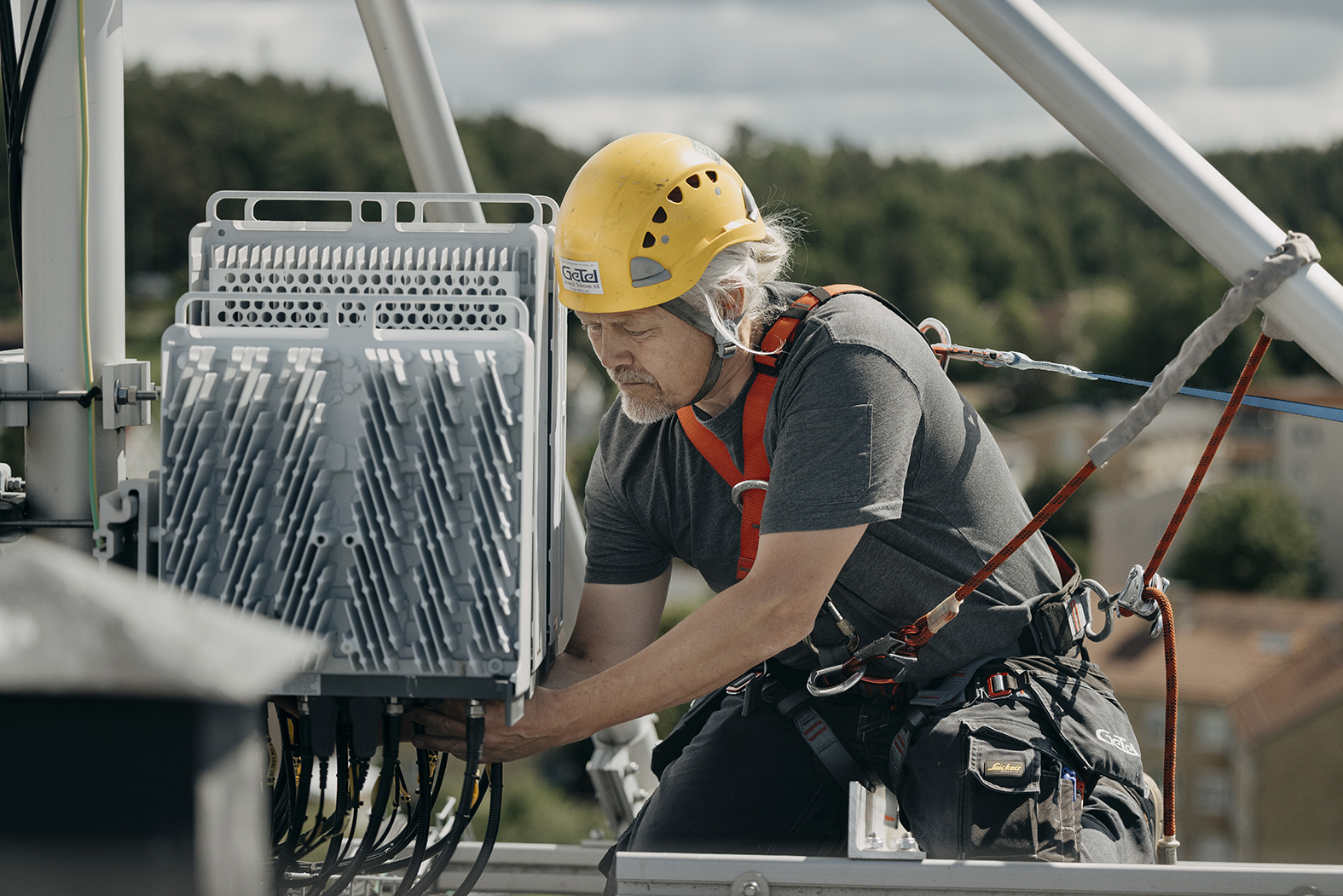 Tekniker i säkerhetsutrustning installerar telekomutrustning på en mast med stad i bakgrunden